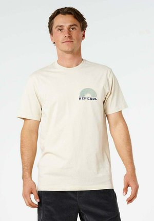 Giovane uomo in piedi che indossa una maglietta beige chiaro con un piccolo logo Rip Curl sul petto sinistro e pantaloni scuri su uno sfondo semplice.