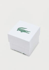 Balts, teksturēts dāvanu iepakojums ar zaļu krokodila logotipu uz vāka un "LACOSTE" izdrukātu zaļā krāsā sānos. Kvadrātveida forma, minimālistisks dizains.