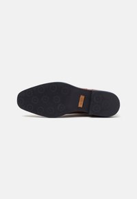 Suela de zapato de goma negra con un patrón de agarre circular y un acento de cuero marrón con un logo en relieve. Acabado liso, diseño antideslizante.