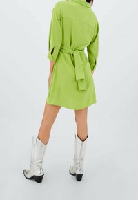 Abito camicia verde con cintura legata, maniche lunghe e spacchi laterali. Indossato con stivali da cowboy argentati lucidi con tacco a blocco.