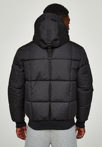 Sort polstret jakke med hætte, der har quiltede sektioner og en detalje med bagrem. Ribstrikkede ærmeafslutninger og linning. Glat ydre tekstur.