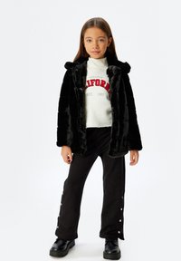Sort faux fur jakke med hætte, udstyret med store knapper; parret med en hvid sweatshirt med rød tekst og sorte flared bukser med trykknapper.