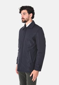 Giacca blu navy con colletto a punta, chiusura con zip, tasche laterali e texture liscia. Presenta una vestibilità su misura e linee pulite.