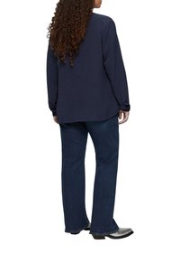 Marineblaues Langarmoberteil mit einem lässigen Schnitt, strukturiertem Stoff und abgerundetem Saum, kombiniert mit dunkelblauen Jeans und glänzenden schwarzen Stiefeln.