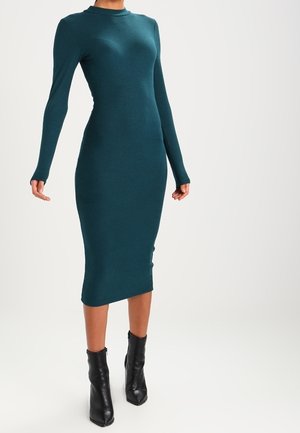 Robe midi en maille côtelée turquoise avec manches longues, encolure ronde et silhouette ajustée. Associée à des bottines noires.