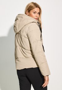 Veste matelassée beige avec une capuche, design quilté et finition texturée. Présente une fermeture éclair noire et un cordon de serrage ajustable sur la capuche.
