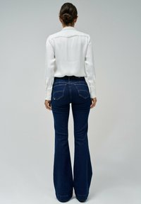 Jeans oscuros acampanados de talle alto, combinados con una blusa blanca de manga larga, vistos desde la parte de atrás, mostrando los detalles de los bolsillos.