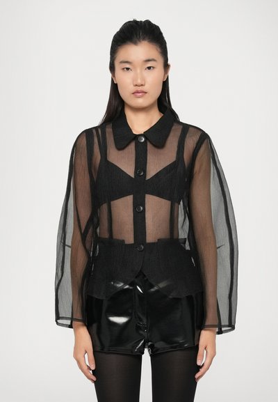 Veste en maille noire transparente avec manches longues translucides, fermeture par boutons, et détails de design structurés portée sur un top noir ajusté et un short.