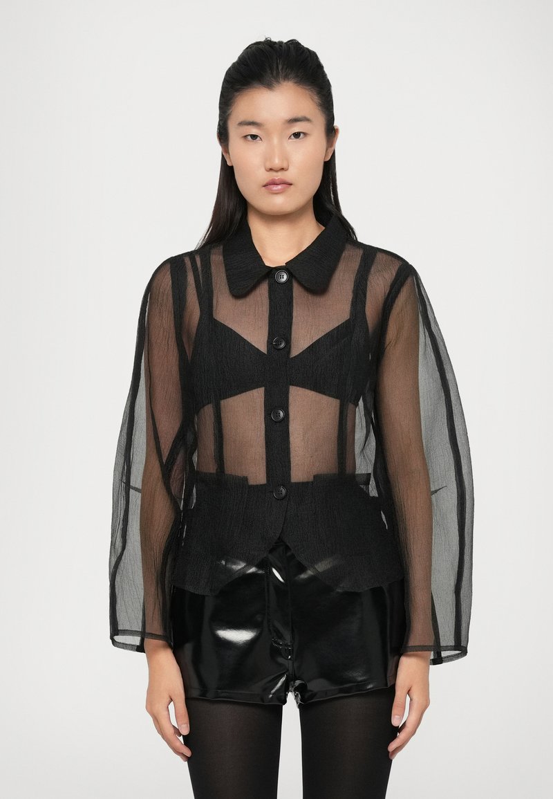 Čierna priesvitná blúzka s golierom, dlhými rukávmi a gombíkovým zapínaním. Obsahuje geometrické výrezy a štruktúrované detaily. Kombinovaná s lesklými šortkami.