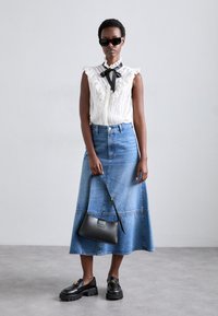 Blusa bianca con rouches e nastro nero, gonna midi in denim blu, borsa nera e mocassini neri chunky su sfondo grigio.