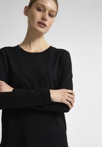 Maglietta nera a maniche lunghe con scollo rotondo, texture morbida e design aderente, con le braccia incrociate, su uno sfondo chiaro.