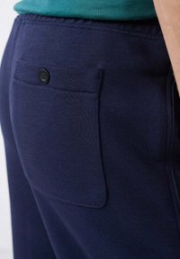 Marineblauwe joggingbroek met een gladde textuur en elastische tailleband. Beschikt over een enkele achterzak met een zwarte knoopsluiting.