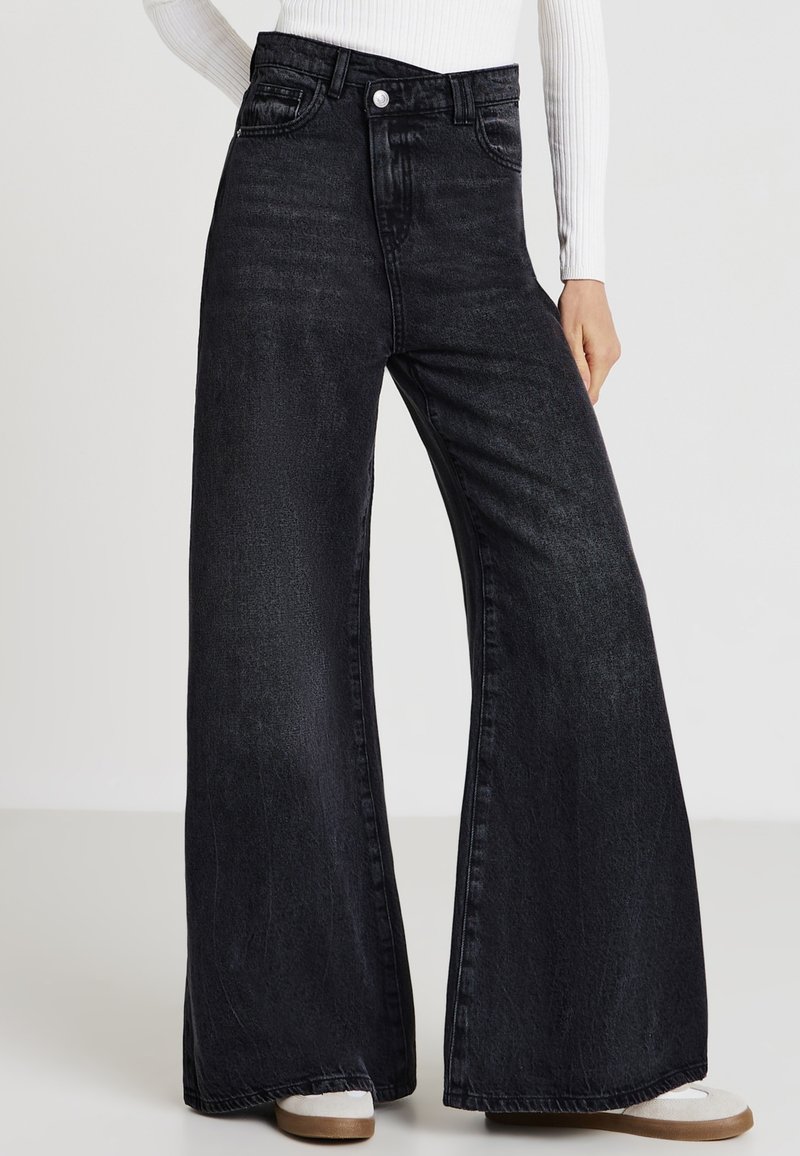 Jean en denim noir à taille haute avec des jambes larges, présentant un design classique à cinq poches et une texture subtile. Le tissu est légèrement usé.