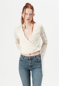 Top blanco de encaje con mangas largas de encaje transparente, escote en V profundo y detalle de lazo en el lateral. Combinado con jeans de mezclilla azul de tiro medio.