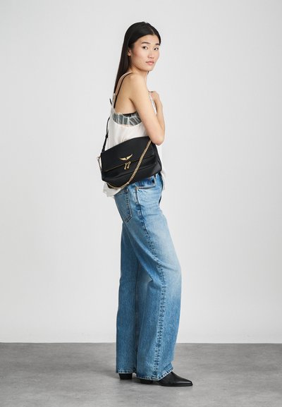 Borsa a spalla in pelle nera con tracolla a catena dorata, tasca con zip e dettagli logo discreti; il modello indossa un outfit casual con jeans a gamba larga.