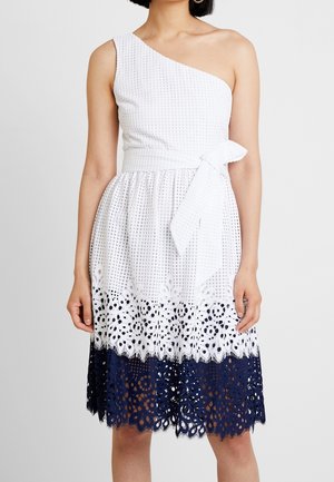 Hvid asymmetrisk kjole med tekstureret stof, taljebånd med sløjfe og nederdel med hulmønster, der har hvide og marineblå lag.