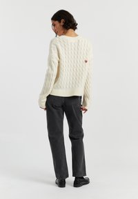 Cremfarbener Kabelstrickpullover mit gerippten Bündchen und Saum, mit einem kleinen roten Herz-Emblem auf dem Rücken; kombiniert mit dunkelgrauen Hosen und schwarzen Schuhen.