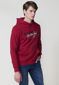 Felpa rossa con tasca frontale, testo in bianco e nero che legge "KODÜCHI Unlimited BRAND." Presenta cordini e polsini a coste.
