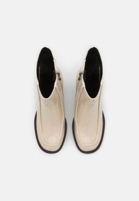 Botines de charol beige con un acabado liso, cremalleras laterales y una suela de goma marrón texturizada, con un diseño de punta redondeada.