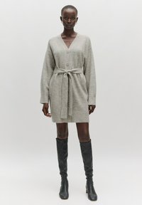 Grijze gebreide cardigan jurk met een V-hals, knopen aan de voorkant en een strikriem om de taille. Kniehoge zwarte aansluitende laarzen maken de look compleet.