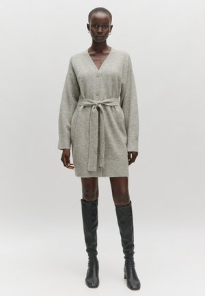 Grijze gebreide cardigan jurk met een V-hals, knopen aan de voorkant en een strikriem om de taille. Kniehoge zwarte aansluitende laarzen maken de look compleet.