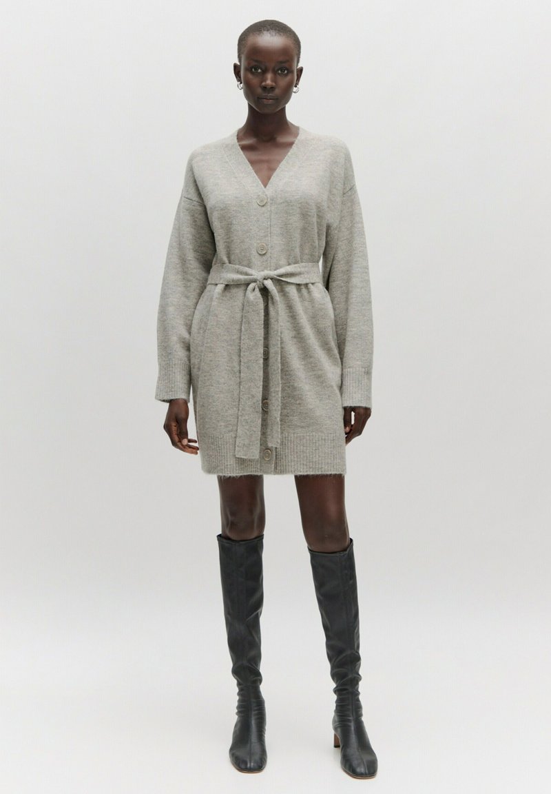 Grijze gebreide cardigan jurk met een V-hals, knopen aan de voorkant en een strikriem om de taille. Kniehoge zwarte aansluitende laarzen maken de look compleet.