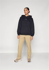 Felpa nera con cappuccio, tasca anteriore e coulisse, abbinata a pantaloni beige e sneakers marrone chiaro, il tutto su uno sfondo uniforme.