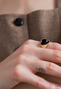 Gold- und schwarze herzförmige Ring mit einem kleinen Diamantakzent, getragen an einer Hand mit einem auffälligen Tattoo, vor einem braunen Stoff-Hintergrund.