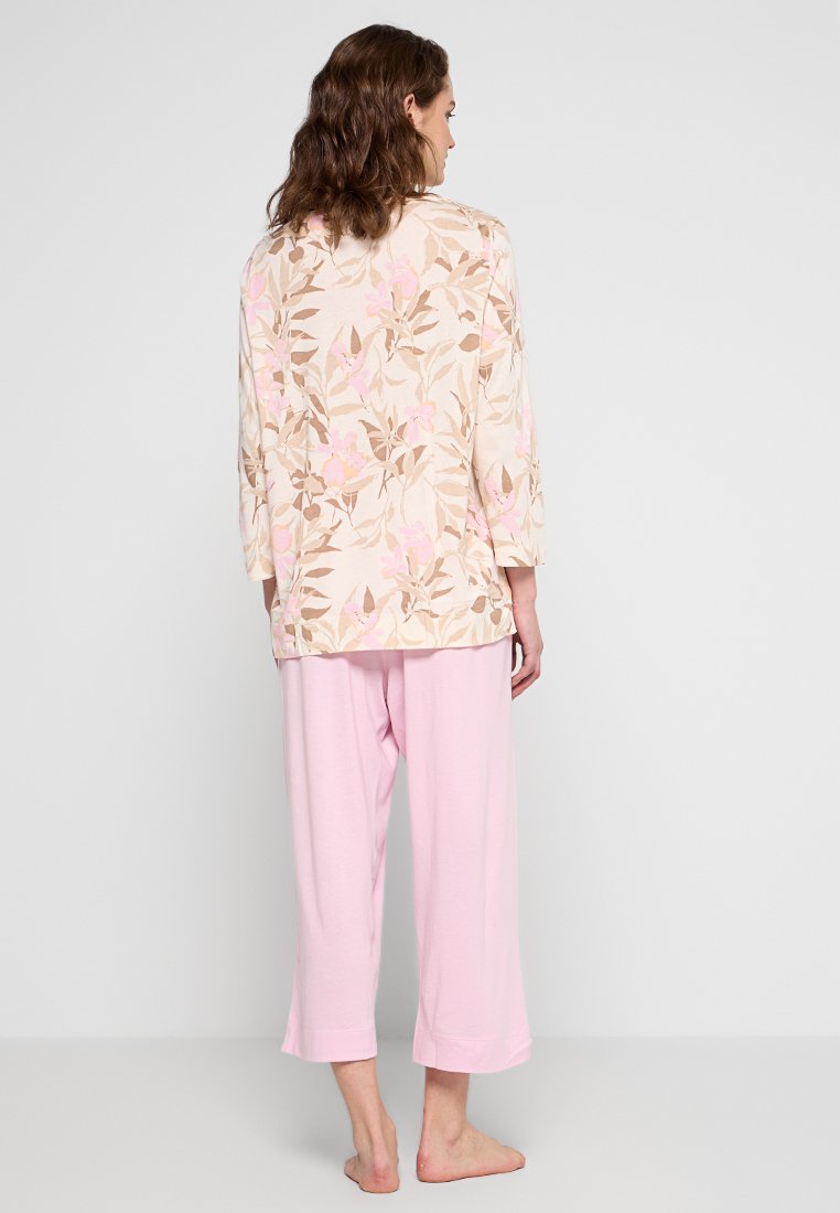 Vrouw die blootsvoets met haar rug naar ons toe staat, gekleed in een crèmekleurige bloemen blouse met lange mouwen en lichtroze wijde broek.