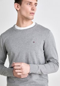 Szary sweter dzianinowy z okrągłym dekoltem, ściągaczami na rękawach i dyskretnym logo na piersi. Miękka struktura, klasyczny krój, uniwersalny design.