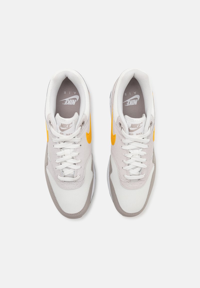 Zapatillas Nike Air Max con parte superior de malla blanca, acentos grises y un swoosh amarillo. Diseño clásico de cordones y cuello acolchado.