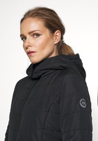 Melnas pūkainās jakas ar kapuci, ar quilted tekstūru, priekšējo rāvējslēdzēju un apaļu logotipu kreisajā piedurknē.
