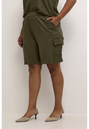 Donna che indossa pantaloni cargo verde oliva fino al ginocchio con tasche, abbinati a décolleté beige con cinturino, in piedi di fronte a uno sfondo bianco.