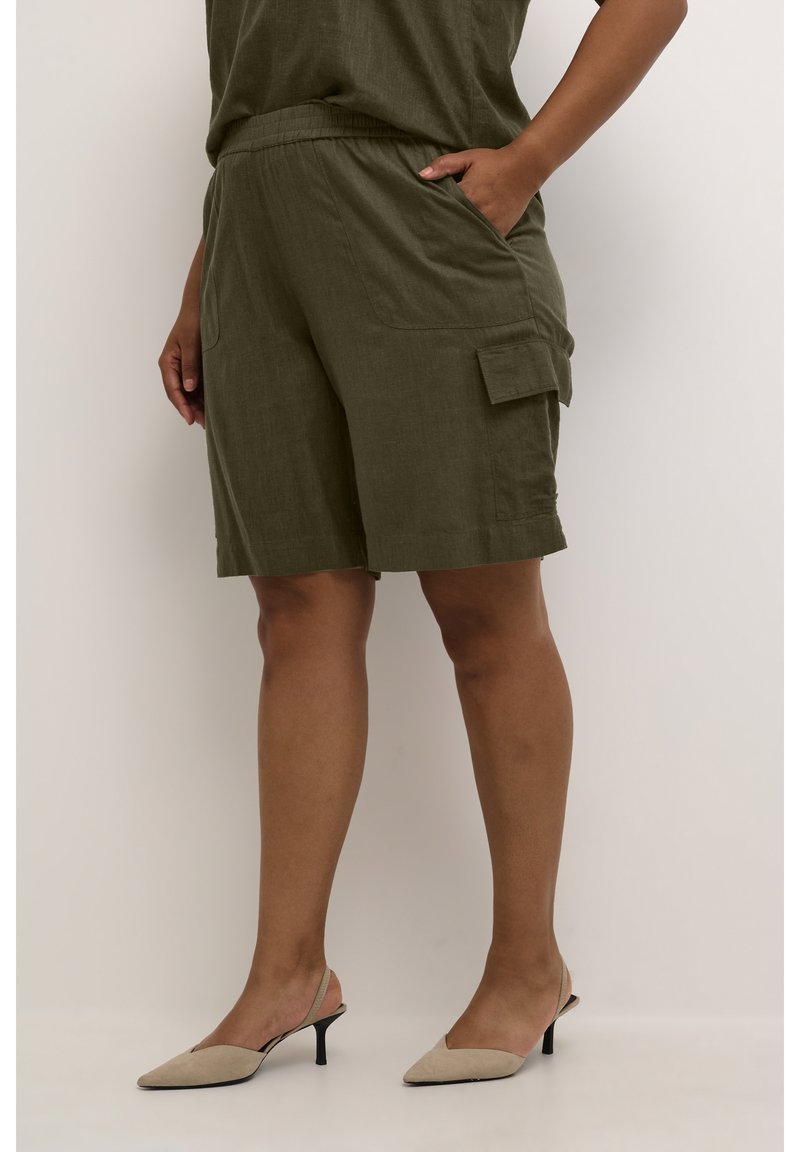 Kvinde iført olivengrønne knælange cargo shorts med lommer, parret med beige slingback sko, stående foran en hvid baggrund.
