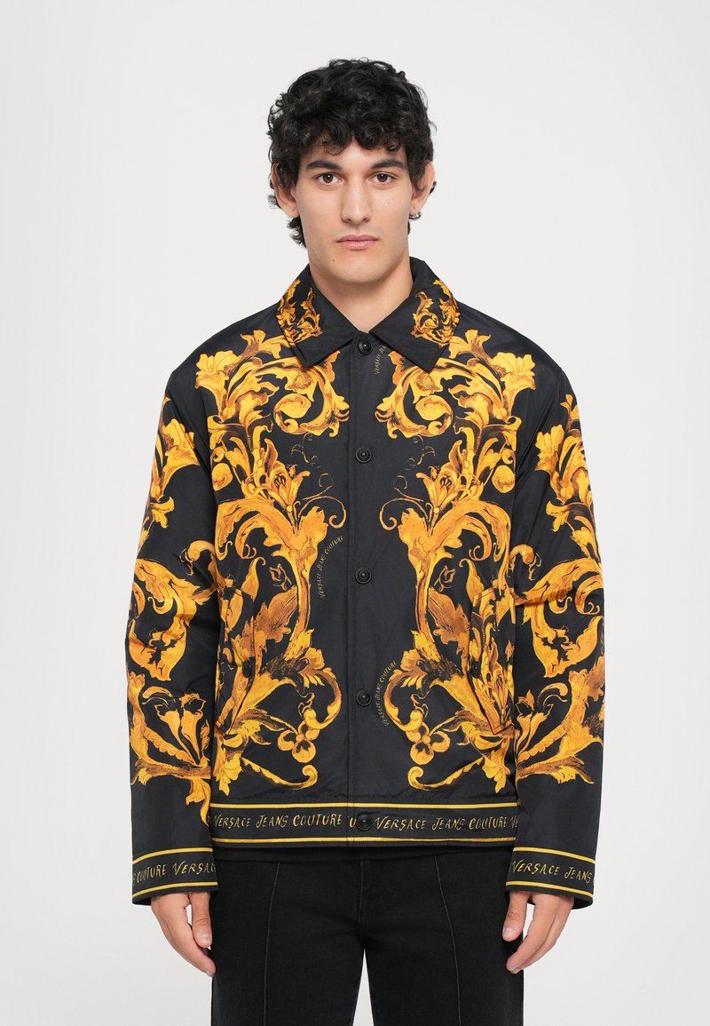 Bărbat îmbrăcat cu o jachetă neagră cu modele florale baroce aurii elaborate și trimuri cu logo-ul Versace Jeans Couture, stând în fața unei fundaluri albe simple.