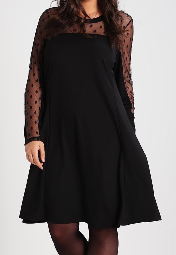 Femme portant une robe noire avec des manches longues noires transparentes à pois et un panneau au niveau de la poitrine, debout devant un fond uni.