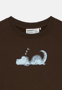 Rjava majica za dojenčke z grafiko modrega dinozavra, ki spi, zaprti oči, z risanimi simboli "zzz" nad glavo.