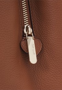 Brązowa skórzana torebka z zapięciem na złoty zamek błyskawiczny i zawieszką w kształcie serca z wygrawerowanym napisem "Kate Spade." Teksturowana powierzchnia.