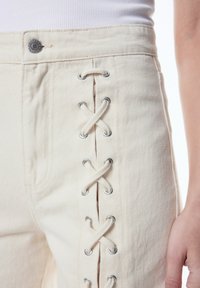 Cremefarbene Denim-Jeans mit Ösen und Schnürdetails entlang der Seite, einem Knopf im Taillenbund und strukturiertem Stoff.