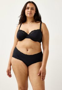 Kvinde med mellemtonet hud og langt mørkt hår iført bralette med sort blondekant og matchende højtaljede trusser, stående mod hvid baggrund.