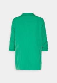 Blazer vert à manches longues avec col et poignets retournés, vu de dos sur un fond blanc uni.