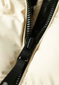 Giacca imbottita beige con zip nera con il marchio "SUPERDRY" e un pratico tirante testurizzato, caratterizzata da un tessuto liscio e cuciture ben definite.
