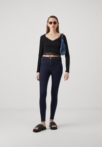 Crop top nero a maniche lunghe con pizzo, jeans skinny a vita alta blu scuro, occhiali da sole rosa e sandali neri. Piccola borsa blu sulla spalla.
