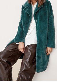 Tealowy futerko z sztucznego włosia z wykładanym kołnierzem, zapięciem na guziki i długimi rękawami, zestawione z brązowymi skórzanymi spodniami o błyszczącej powierzchni.