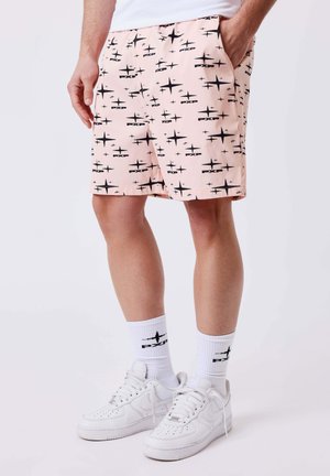 Uomo con pantaloncini rosa a motivi di stelle nere, calzini bianchi alti con logo nero e scarpe da ginnastica bianche, in piedi contro uno sfondo semplice.