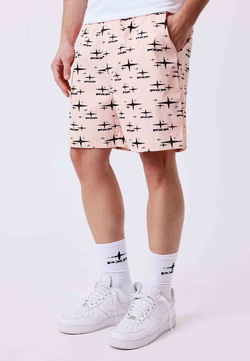 Homme portant un short rose orné de motifs d'étoiles noires, des chaussettes basses blanches avec un logo noir et des baskets blanches, debout devant un fond uni.