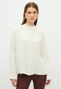 Witte blouse met lange mouwen en mandarinnekraag, ontspannen pasvorm, gladde textuur en knoopsluiting aan de voorkant. Gecombineerd met bruin aansluitende broek.