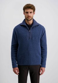 Strickpullover aus strukturiertem blauen Stoff, mit halbem Reißverschluss, gerippten Bündchen und Saum. Getragen mit dunklen Hosen.