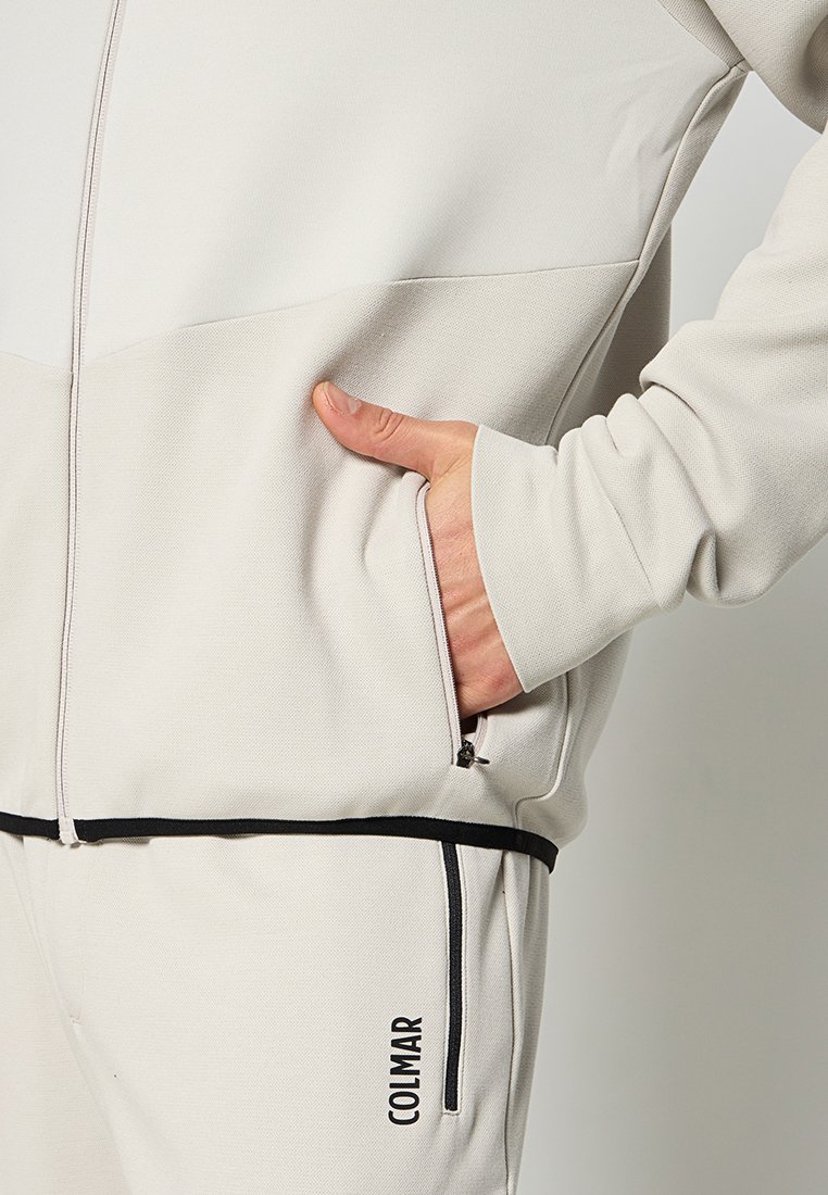 Mano nella tasca con zip di un giubbotto beige chiaro abbinato a pantaloni coordinati, caratterizzati da un logo Colmar nero e dettagli in nero.