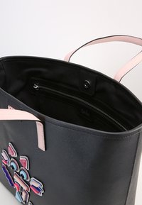 Svart tote-väska med rosa handtag, prydd med färgglada geometriska lappar framtill och ett innerfack med dragkedja och tryckknappslås.